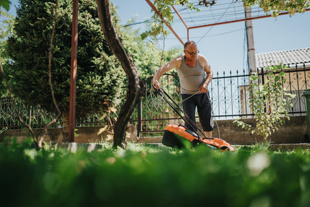 Man mowing a sunny backyard lawn with an orange lawn mower in a residential garden