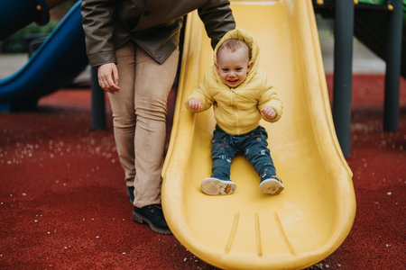 Parent guiding a toddler on a yellow slide at an outdoor playgroundの写真素材