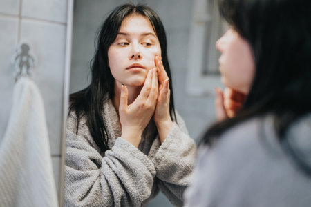 Young woman applying skincare routine while reflecting on her image in mirrorの写真素材
