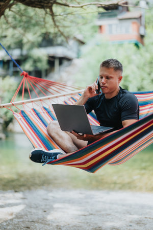 Man working remotely with a laptop and phone in a colorful hammock outdoorsの写真素材