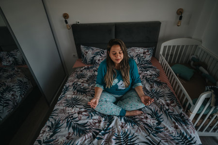 Woman meditates on a bed in a cozy bedroom beside a baby crib with floral sheets