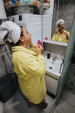 Woman in yellow robe applying makeup in bathroom, smiling at mirror during morning routine