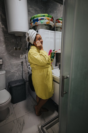 Woman in yellow bathrobe applying skincare in laundry room by washing machineの写真素材