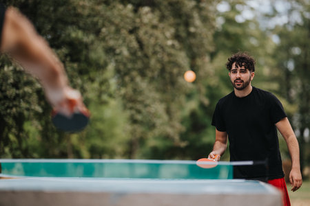 Young man in black shirt plays table tennis with a friend outdoors in a park settingの写真素材