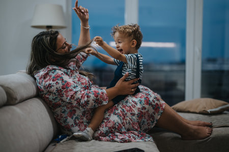 Mother and young son playfully laugh together on a floral couch at homeの写真素材