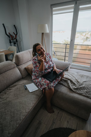 Woman working from home on a cozy sofa, talking on the phone while using a laptopの写真素材