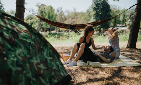 Friends relax on a picnic blanket in a park, sharing laughter near hammock and tentの写真素材