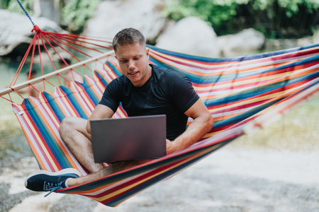 Man working remotely on a laptop while relaxing in a colorful hammock outdoorsの写真素材