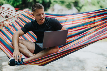 Man working on a laptop while sitting in a colorful outdoor hammockの写真素材