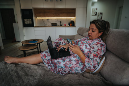 Pregnant woman relaxing at home with laptop and phone in a cozy modern living spaceの写真素材
