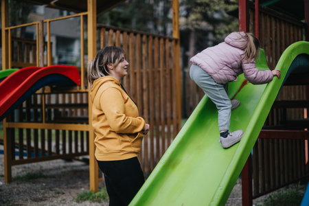 Mother watching her daughter climbing playground slide in sunny city parkの写真素材