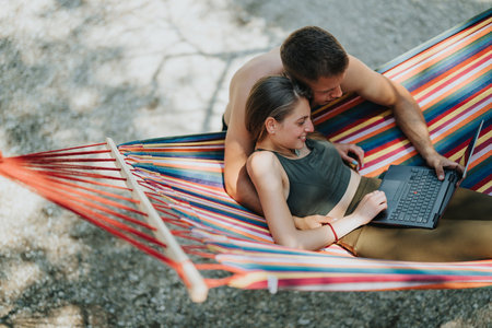 Couple relaxing on colorful hammock while working remotely on a laptop outdoorsの写真素材