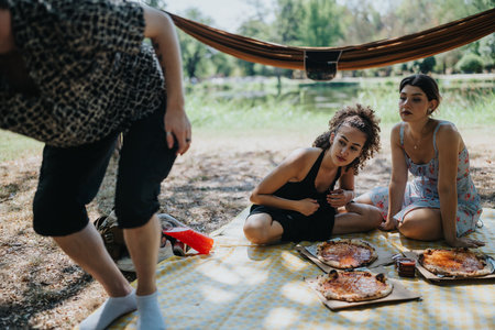 Friends enjoy a casual outdoor picnic with pizza on a checkered blanket under a hammockの写真素材