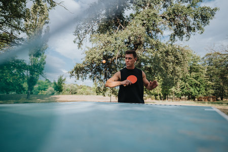 Man playing table tennis outdoors in a park, focused on his serve under trees on a sunny dayの写真素材