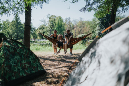 Friends relax in a hammock at a lakeside campsite with tents and trees in viewの写真素材