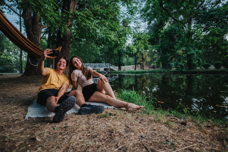 Two friends take a sunny selfie by a lake in a lush park, relaxing togetherの写真素材