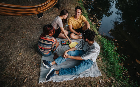 Friends enjoy a camping chill by the river, sharing a board game on a blanket outdoorsの写真素材