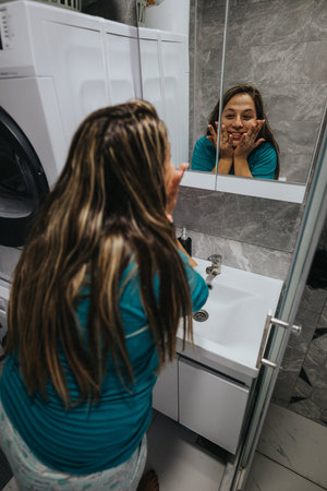 Woman in teal shirt smiles at her reflection in a bathroom mirror, hands on face, cheerful momentの写真素材