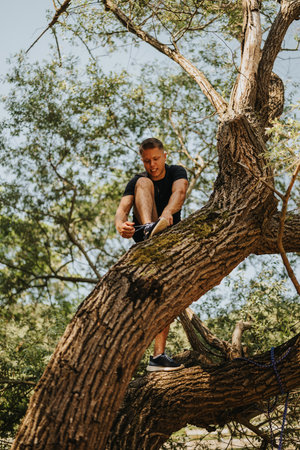 Man sitting on a tree branch outdoors tying his shoelaces