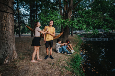 Friends laugh and relax in a park by a hammock near a pond and wooden bridgeの写真素材