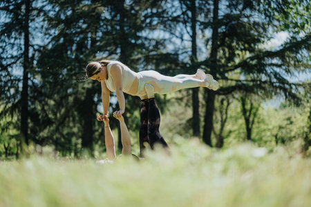 Two individuals practicing partner yoga in a sunny park with tall trees and lush greenery, balancing and engaging in an acro yoga pose.の写真素材