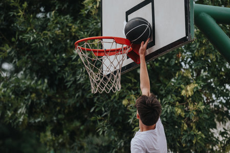 Young man performs a slam dunk on an outdoor basketball hoop at a park.