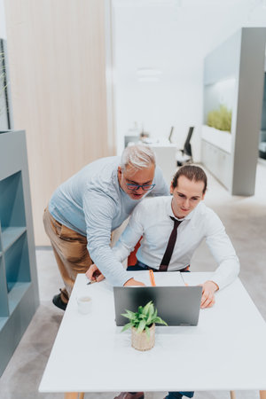 Mentoring moment in a modern office as a senior colleague guides a younger coworker at the laptopの写真素材
