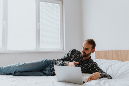 Man Relaxing on Bed While Using Laptop in Bright Roomの写真素材