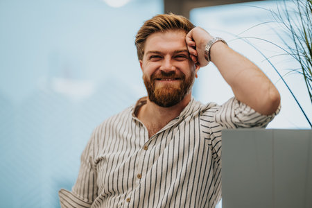 Smiling man in striped shirt leans on office desk, conveying relaxed confidence and friendly professionalism.の写真素材