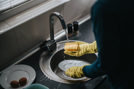 Person wearing yellow gloves washing a white plate under running water in a kitchen sink, emphasizing cleanliness and hygiene.の写真素材