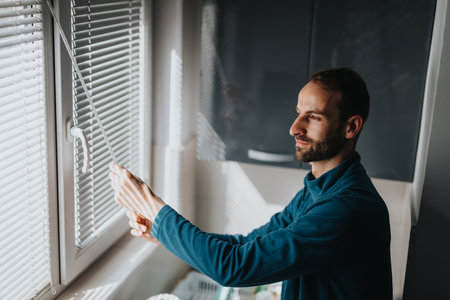 Man adjusting window blinds in a sunlit modern home interiorの写真素材