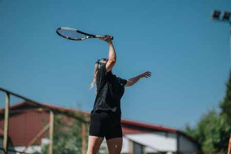 Female tennis player serving ball during outdoor match under clear blue sky on a sunny afternoonの写真素材