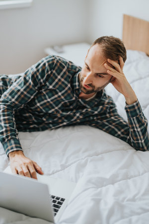 Man in plaid shirt using laptop while relaxing on a comfortable bedの写真素材