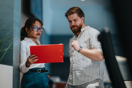Two professionals discuss a red notebook during a team collaboration in a modern office meeting.の写真素材