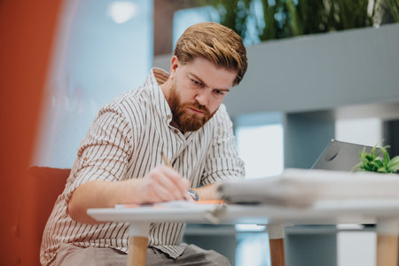 Focused man with striped shirt writes at a white desk in a bright modern office