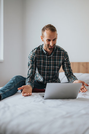 Smiling man using a laptop while sitting on a comfortable white bedの写真素材