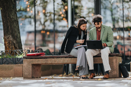 Two colleagues review documents and a laptop on a park bench in an urban city settingの写真素材