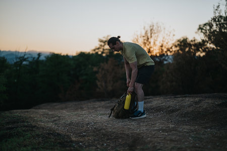 Man prepares hiking gear at dusk on a trail, packing a backpack and water bottle