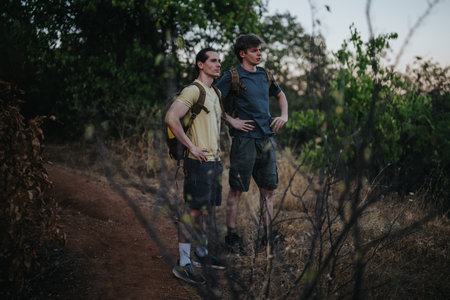 Two hikers with backpacks stand on a dirt trail in a forested area, ready for adventure