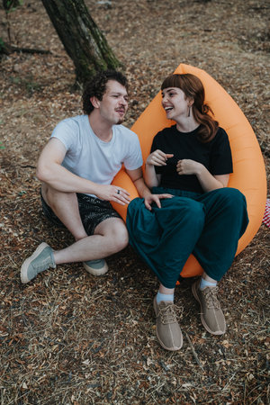 Two friends laugh and chat while relaxing on an orange camping chair in the forest