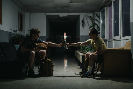 Two friends share a drink in a dim hallway, sitting apart with backpacks and plants nearby