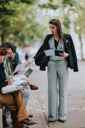 Businesswoman in gray suit distributes papers to a group outdoors in a city setting