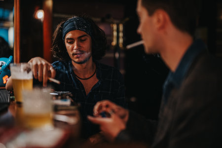 Friends at a dim bar sharing drinks and a cigarette during a moody late-night chatの写真素材