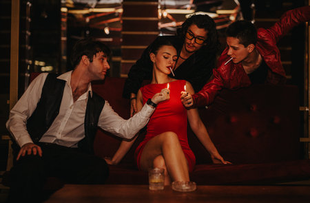 Group of friends in a chic night club sharing cigarettes, with a woman in a red dress seated on a velvet sofaの写真素材