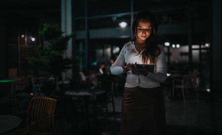 Woman at night in a dim cafe using a tablet, focused and calm under soft lightの写真素材