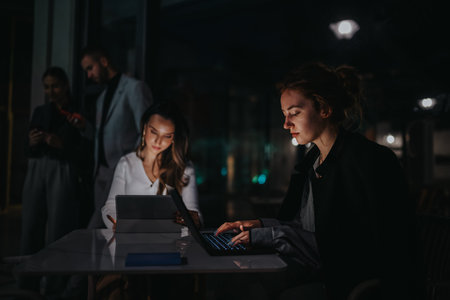 Evening office team collaborates on laptops and tablets in a modern dark workspaceの写真素材