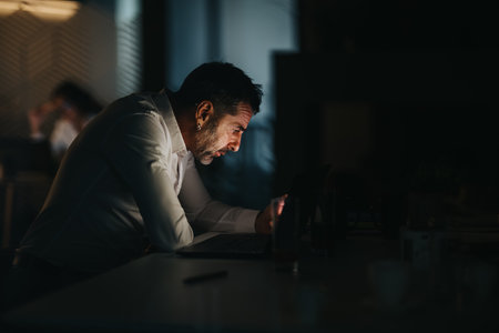 Focused man working late at night in an office, typing on a laptop beside a glassの写真素材