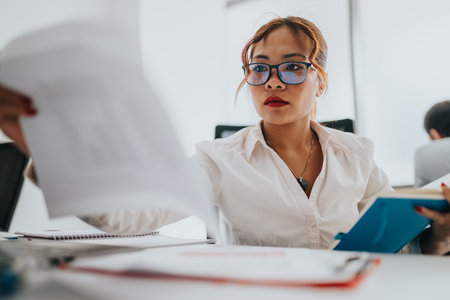 Focused businesswoman reviews documents in a bright office, holding a blue folder and a tabletの写真素材