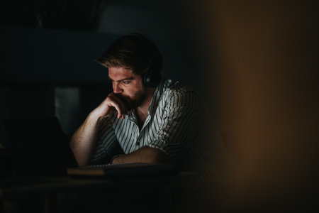 A thoughtful man with headphones works late at night in a dimly lit office