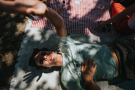 Friends enjoying a sunny outdoor picnic and taking a group selfie on a blanketの写真素材